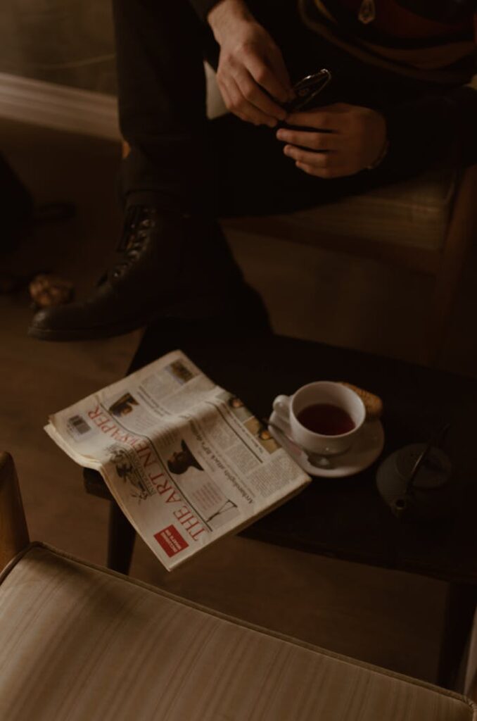 From above of crop anonymous man with eyeglasses in hands sitting on chair near small table with newspaper and tea set during breakfast